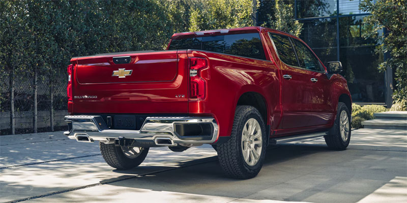 A rear-angle view of a cherry red Chevrolet Silverado LTZ parked on a concrete driveway with a modern building and green trees in the background.